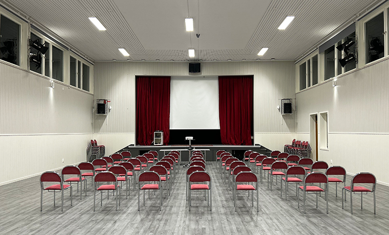 En stor ljusgrå sal full av röda stolar som är vända mot en scen med röd ridå